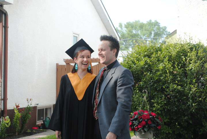photo of girl in grad cap and gown standing with her father both are laughing as they stand in front of bushes and flowers